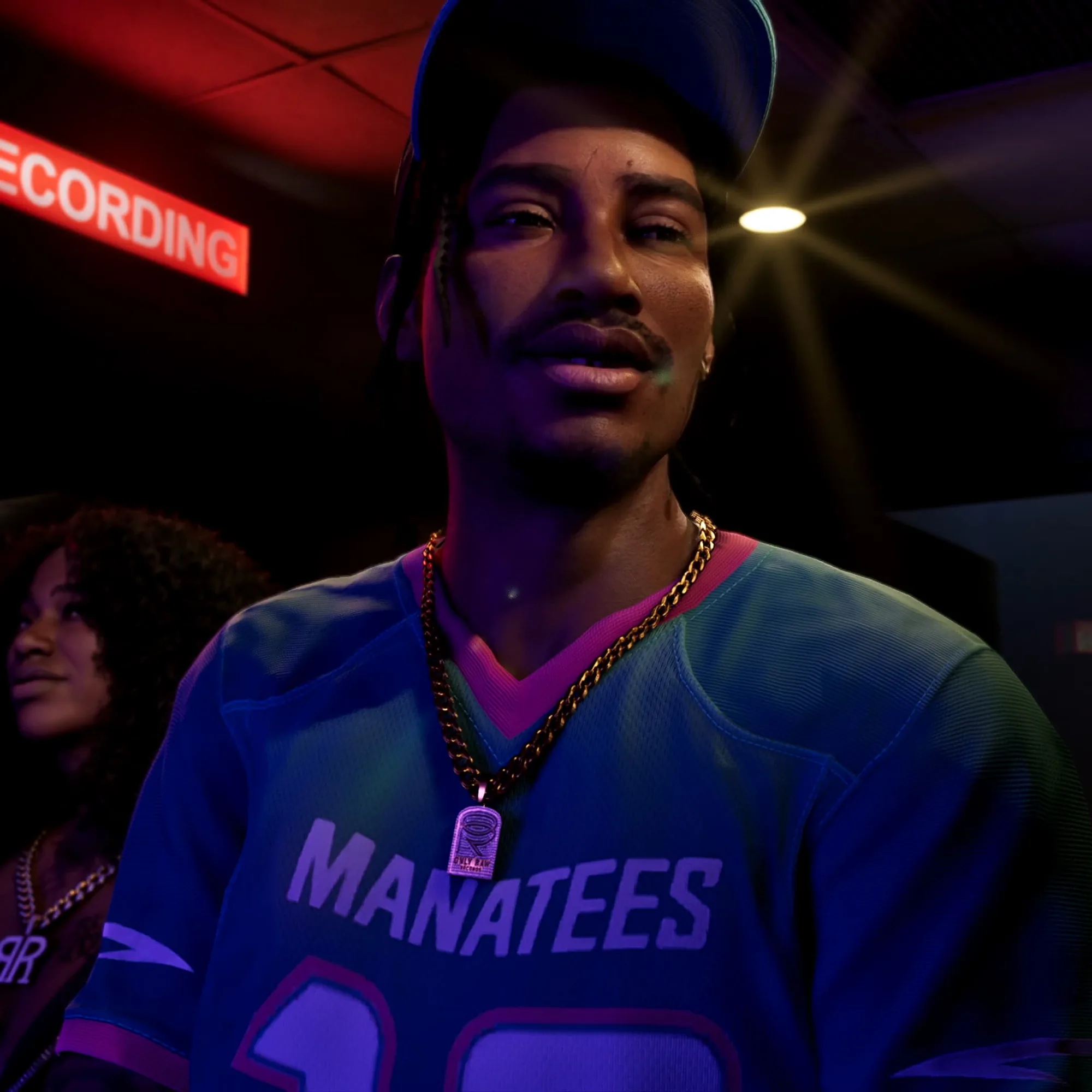 A man in a blue Manatees jersey, cap and gold chain necklace stands in a venue lit by purple, blue and red stage lights.
