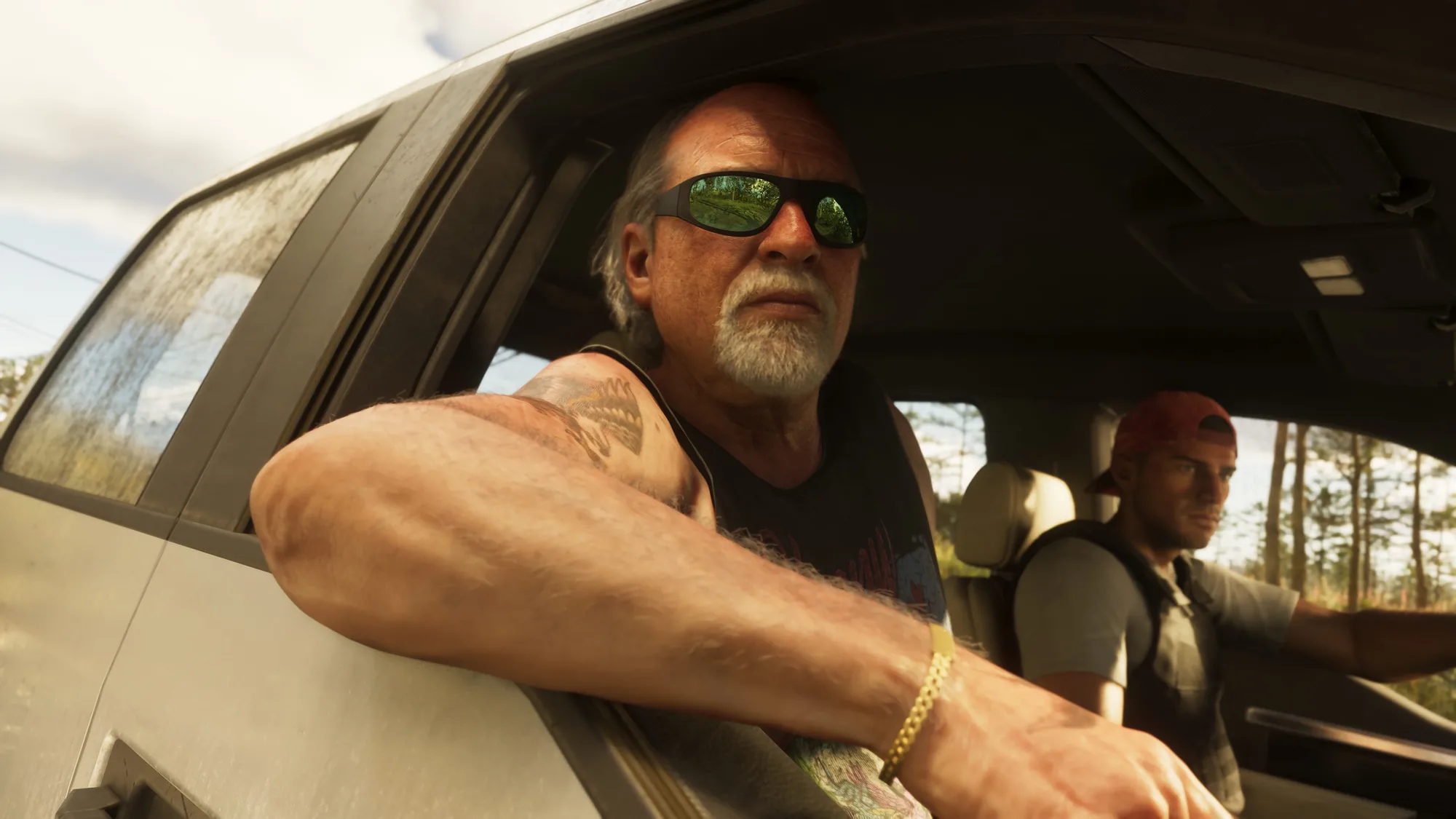 A middle-aged man in dark sunglasses and a black sleeveless shirt leans out of the window of a light-coloured vehicle on a rural road lined with pine trees, another passenger beside him.