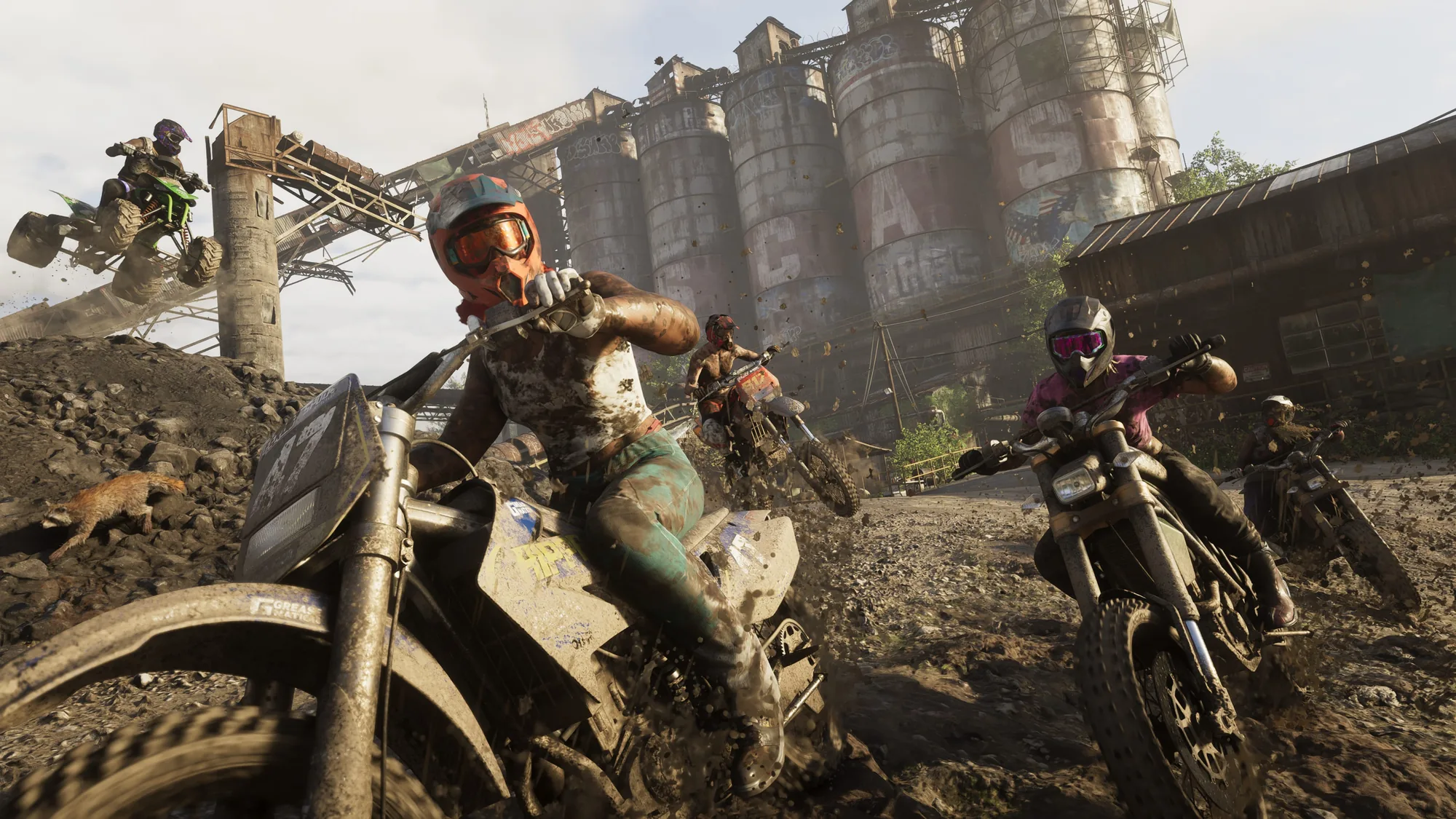 Mud-covered riders on dirt bikes and motorcycles gather in a dusty industrial yard beneath towering grain silos in daylight.